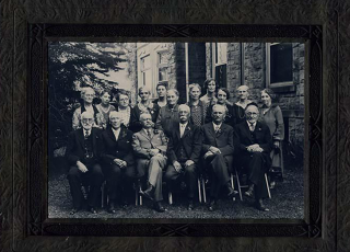 Black-and-white group portrait of the first University of Utah faculty, from the Toronto, Joseph B. Photograph Collection. About 18 men and women pose outdoors beside a stone building, with the men seated in front in suits and the women standing behind in dresses. The photo is mounted in a dark embossed frame.